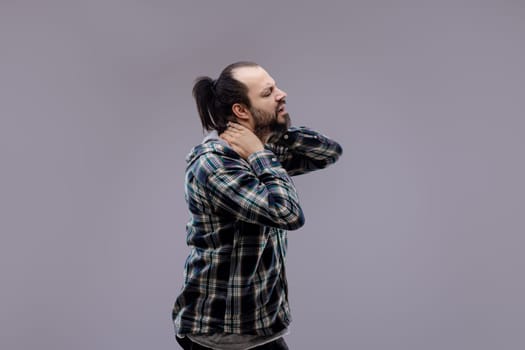 Casual man with beard and ponytail wearing a plaid shirt, standing and suffering from neck pain, massaging his neck with a painful expression