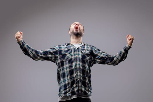 Bearded man celebrates victory with raised fists, showing pure joy and excitement against a gray studio background