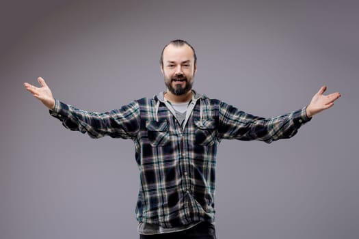 Bearded man stands with arms wide open, smiling happily in a casual shirt against a grey studio background, radiating positivity and confidence