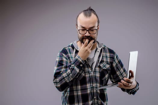 Focused web developer with laptop contemplates coding problem in studio. Gray background enhances serious atmosphere. Expertise and determination evident