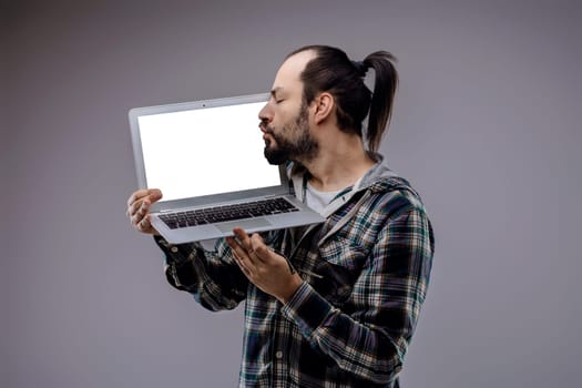 Passionate programmer kisses his laptop screen, showing love for coding. With long hair tied back, he wears a casual shirt against a gray background. His focus on technology and success is clear