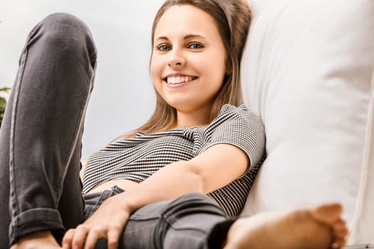 Young woman sits comfortably on a white couch at home, relaxing with a smile, embodying wellbeing in a candid moment of joy