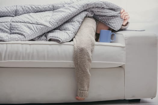 Tired woman rests on the sofa with a hot drink, feeling sick and seeking comfort at home