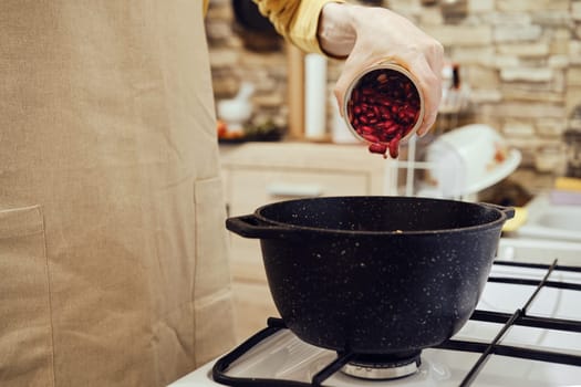 Unrecognizable man pours canned beans from tin can into a saucepan on a gas stove
