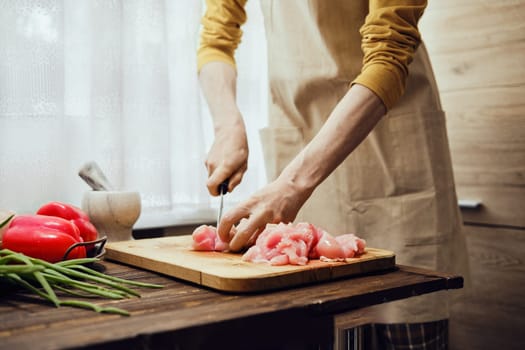 Unrecognizable man in apron slicing raw chicken fillet with kitchen knife in the kitchen