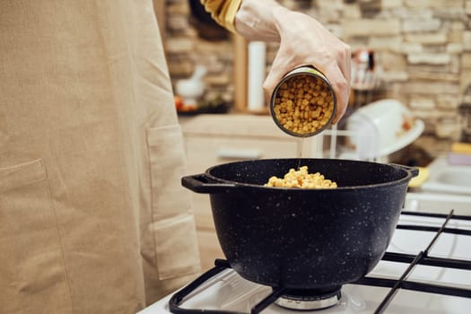 Unrecognizable man pours canned corn from tin can into a saucepan on a gas stove