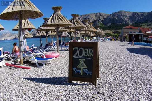 Beach scene with teepee,sun umbrella and beach loungers in front of the beautiful mountain scenery of Baska/island of Krk - 20 Euro/day for the deck chair is not cheap  Theme picture Croatia / Hrvatska / Croatia 2024