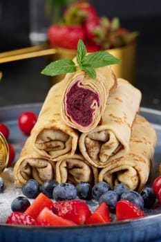 Crepes stacked in a pile, one of which is cut and filled with berry filling. The plate is also decorated with blueberries, strawberries, and cherries