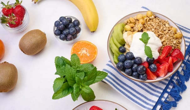 Granola with strawberries, kiwi, banana and blueberries in a round plate on a white table. Healthy and tasty food, top view
