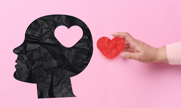 A black silhouette head, with a heart cut out in the area where the brain is, creating a heart-shaped empty space. The hand is holding a red paper heart, about to insert it into the cut out heart in the head.