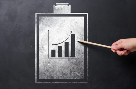 A folder holder for sheets of paper with a graph drawn with white chalk on a black chalk board, a hand with a pencil indicates the growth of indicators