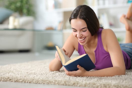 Happy woman laughing reading a paper book lying on a carpet at home