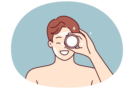 Man demonstrates jar of cream bought in cosmetic store to moisturize his face or get rid of wrinkles. Young guy with bare shoulders recommends purchasing nourishing balm or cream for skin care