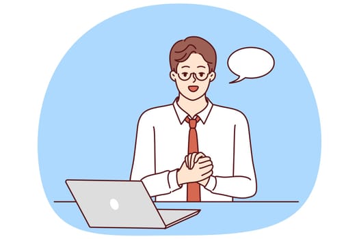 Confident businessman is sitting at table with laptop and looking at camera recording business training. Guy in business clothes with dialogue cloud works as teacher online training courses