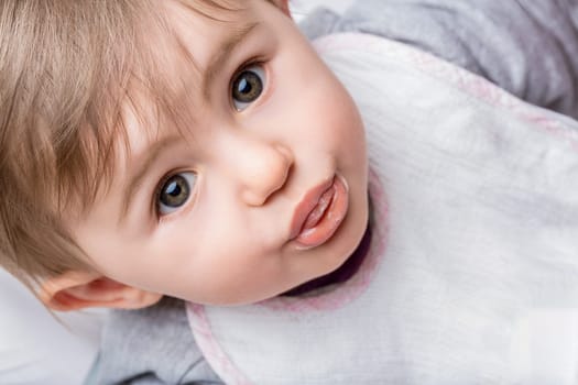 Sweet baby girl in a gray shirt and white bib makes a funny face with her mouth wide open, exuding innocence and joy