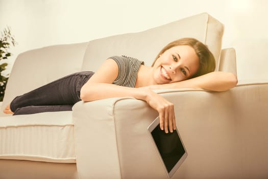 Young woman relaxes on her couch at home, smiling and browsing on her tablet, enjoying leisure time