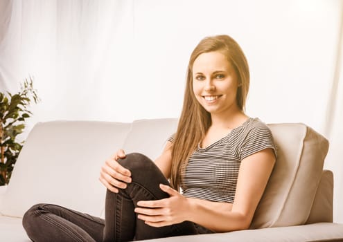 Young woman sits happily on her sofa, embracing her knees in a bright, clean home. She looks relaxed, content, and enjoys a quiet moment alone in her cozy living room, basking in natural light