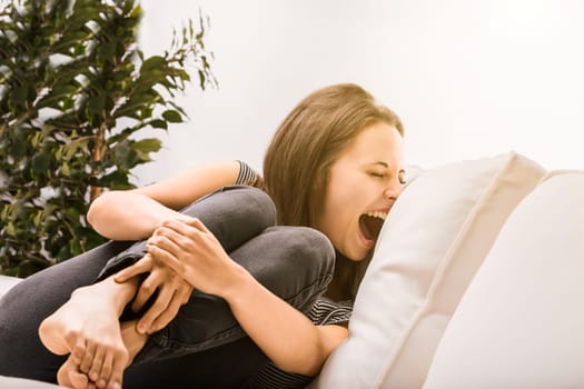 Stressed woman on white sofa screams at home, overwhelmed by anxiety and burnout, surrounded by plants and natural light