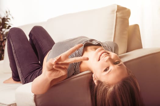 Young woman is lying upside down on her couch, smiling and gesturing a peace sign. She is enjoying her free time at home
