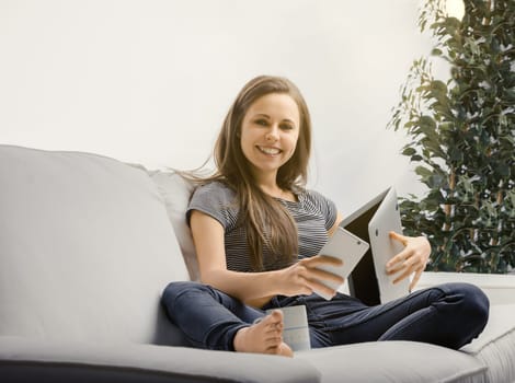 Young woman is sitting on her sofa using a laptop and digital tablet while enjoying a cup of coffee. She is smiling and appears to be enjoying working from home