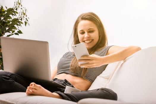 Young woman is spending time at home, relaxing on her couch while using her laptop and smartphone. She is enjoying her downtime by catching up with friends and family online