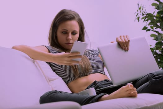 Young woman is lying on her couch, holding her laptop and browsing the internet on her smartphone. She has a neutral expression