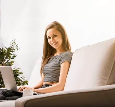 Young woman smiling while working on her laptop, sitting on a comfortable sofa in her living room, enjoying the comfort and independence of working remotely