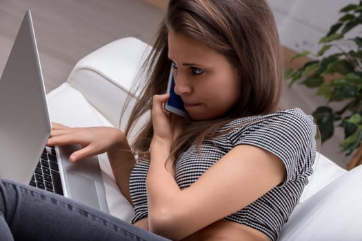 Stressed young woman multitasking on a sofa with laptop and phone, overwhelmed by remote work pressures