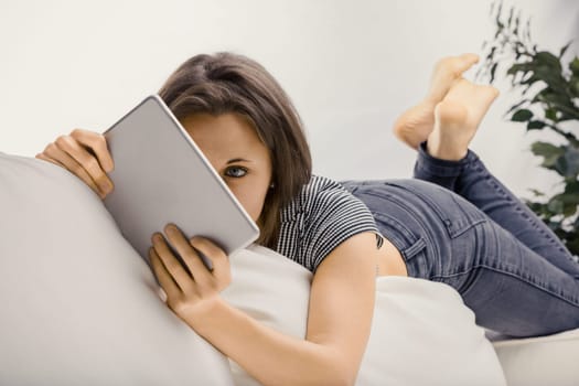 Young woman enjoying her digital tablet on a white couch at home, browsing, streaming, and connecting online in a casual and relaxed manner