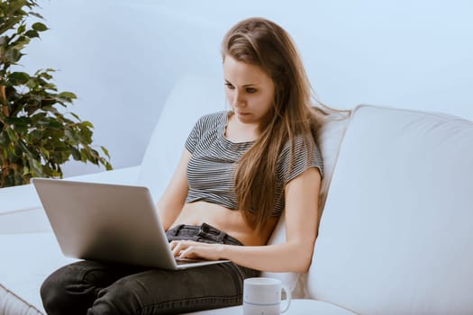 Young woman works comfortably on a laptop in her modern living room, blending work and relaxation seamlessly at home