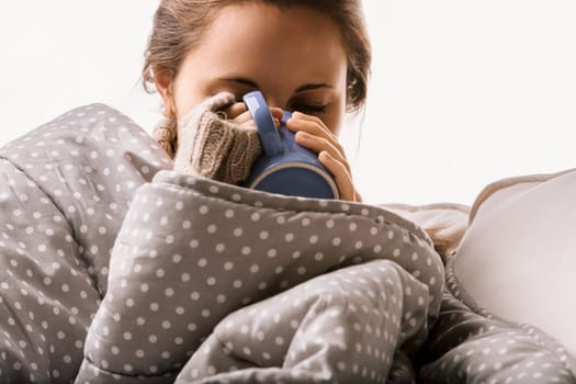 Tired woman snuggles under a blanket, sipping a hot drink for comfort at home. She seeks rest and recovery in her living room during winter