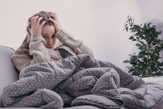 Young woman sits on a cozy sofa at home, wrapped in a warm blanket, looking sad and worried, reflecting feelings of unhappiness and anxiety