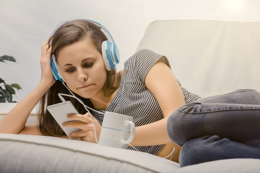 Young woman is lying on her couch, listening to music and looking at her phone with a bored expression. She is holding a mug in her other hand