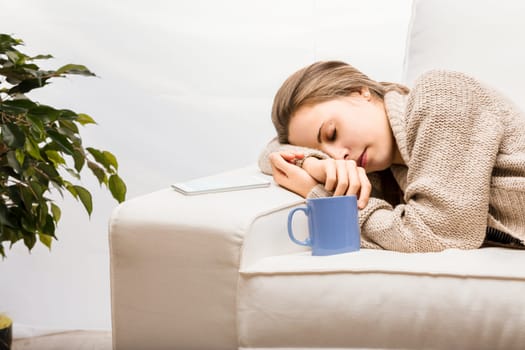 Young woman is sleeping on a comfortable sofa in her living room, with a cup of herbal tea and her smartphone next to her on the sofa