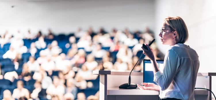 Female speaker giving a talk on corporate business conference. Unrecognizable people in audience at conference hall. Business and Entrepreneurship event