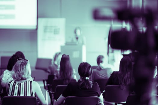 Business and entrepreneurship symposium. Female speaker giving a talk at business meeting. Audience in conference hall. Rear view of unrecognized participant in audience.