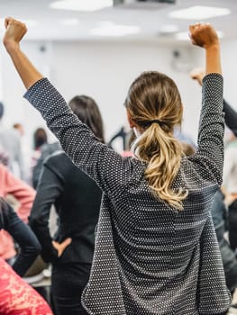 Life coaching symposium. Speaker giving interactive motivational speech at business workshop. Rear view of unrecognizable participants feeling empowered and motivated, hands raised high in air.
