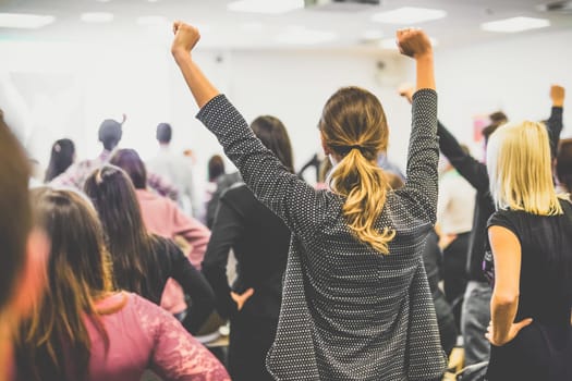 Life coaching symposium. Speaker giving interactive motivational speech at business workshop. Rear view of unrecognizable participants feeling empowered and motivated, hands raised high in air.