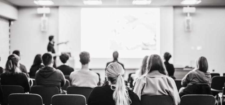 Speaker giving a talk in conference hall at business event. Rear view of unrecognizable people in audience at the conference hall. Business and entrepreneurship concept. Blue tones black and white image