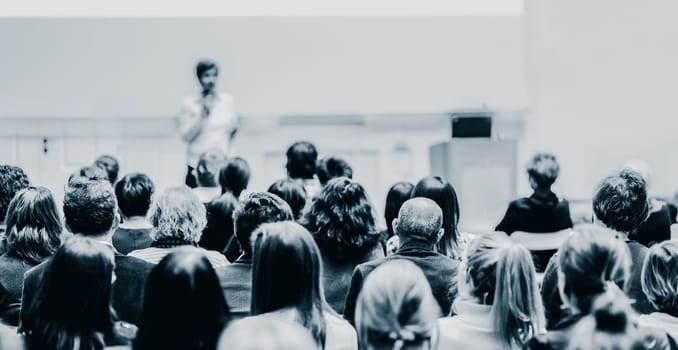 Female speaker giving presentation in lecture hall at university workshop. Audience in the conference hall. Rear view of unrecognized participant in audience. Scientific conference event.