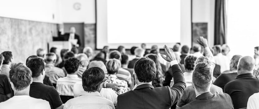 I have a question. Group of business people sitting at the chairs in conference hall. Businessman raising his arm. Conference and Presentation. Business and Entrepreneurship. Black and white.