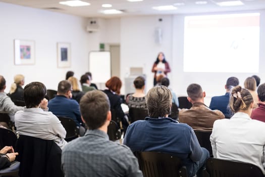 Business and entrepreneurship symposium. Female speaker giving a talk at business meeting. Audience in conference hall. Rear view of unrecognized participant in audience. Copy space on whitescreen.