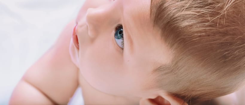 Close up portrait of a beautiful baby boy with blue eyes lying on his back on a white blanket, looking up with curiosity