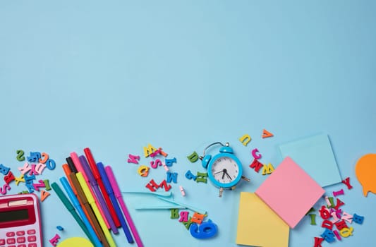 Stationery laid out on a blue background. Multi-colored felt-tip pens, calculator, alarm clock, sticky notes and wooden letters, preparing for study or work, organizing workspace