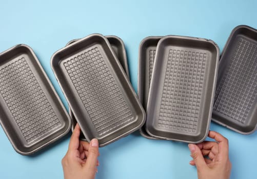 Hand holds black disposable polystyrene foam rectangular plate for packaging vegetables and meat on a blue background, top view