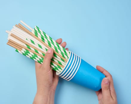 Female hand holding paper cocktail straws and paper cups on a blue background, top view