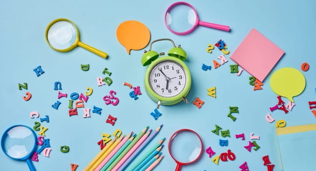 Stationery laid out on a blue background. Multi-colored felt-tip pens, alarm clock, sticky notes and wooden letters, preparing for study or work, organizing workspace