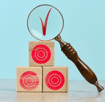 Wooden cubes with red targets stacked on top of each other. On the top cube there is a magnifying glass, through which you can see the red check mark, achieving the goal and checking the results