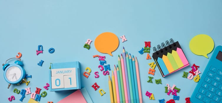 Stationery laid out on a blue background. Multi-colored felt-tip pens, calculator, alarm clock, sticky notes and wooden letters, preparing for study or work, organizing workspace