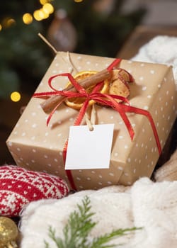 Gift tag on a Christmas present near cosy heart and sweater against Christmas tree lights, close up, copy space. Warm winter family atmospheric mock up, magical Holiday time